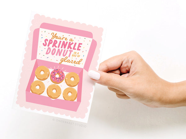 A hand holds the Youre a Sprinkle Donut Greeting Card by Onderkast Studio, featuring a pink box with five plain donuts and one sprinkled donut. The card reads: You’re a sprinkle donut in a box of glazed.