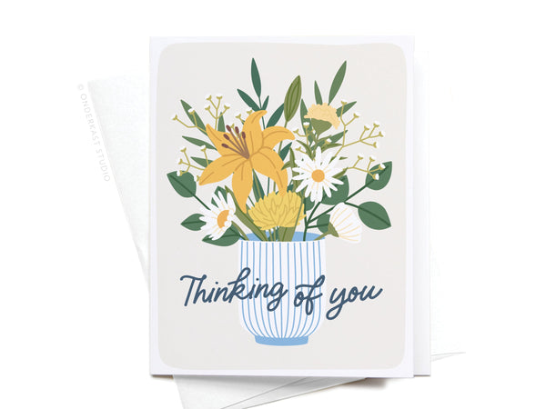 The Onderkast Studio “Thinking of You Flowers” Greeting Card features a charming floral design with yellow lilies, daisies, and greenery in a blue and white vase. “Thinking of You” is elegantly scripted below the arrangement.