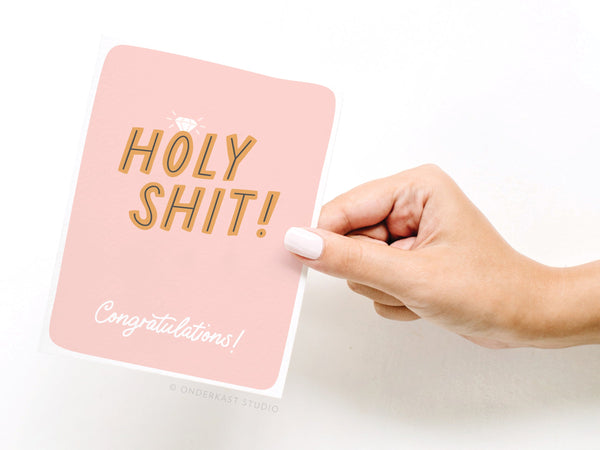 A hand with white nail polish holds the Onderkast Studio Holy Shit Engagement Ring Greeting Card, featuring bold HOLY SHIT! and a script Congratulations! below.