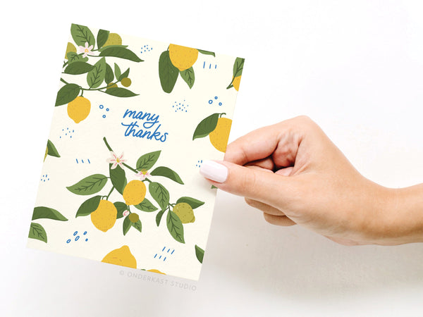 A hand with pale pink nails holds the Onderkast Studio Many Thanks Lemon Pattern Greeting Card, featuring illustrated lemons, green leaves, and white flowers. Many thanks appears in blue cursive on the front.